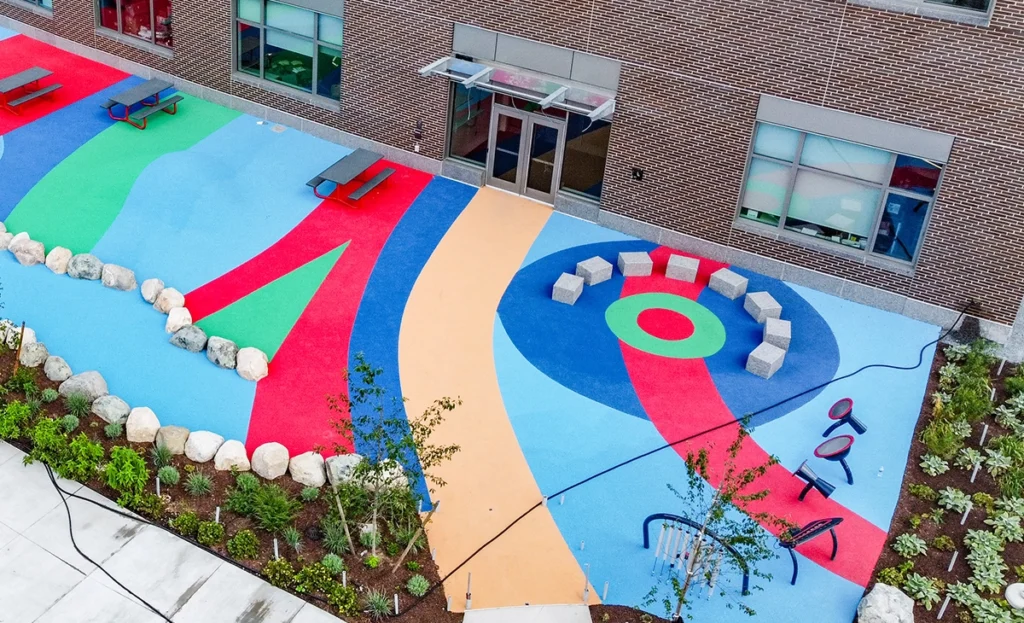 A colorful outdoor playground with abstract patterns, benches, stone blocks arranged in a circle, playground equipment, and landscaped greenery next to a brick building.