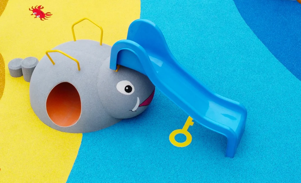 A playground slide shaped like a blue and gray animal with yellow handles sits on a colorful rubber surface with a small red crab illustration nearby.