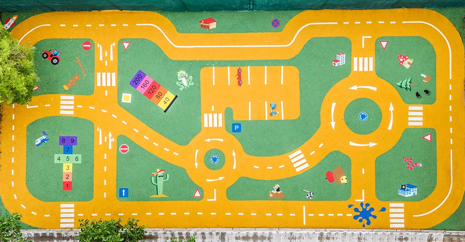 A colorful playground mat features a winding road track with intersections, parking spaces, traffic signs, and playful illustrations like animals, vehicles, and hopscotch squares.