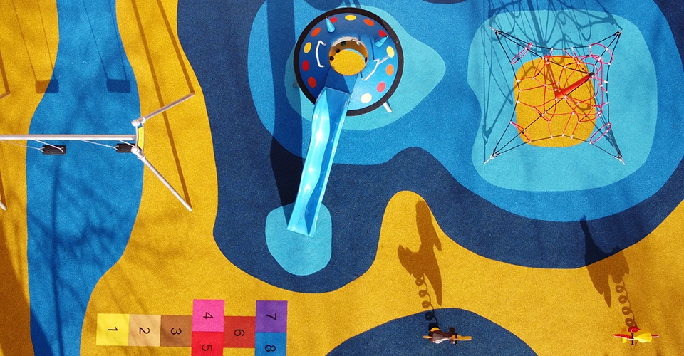 Aerial view of a colorful playground with a slide, climbing net, hopscotch, and two children casting long shadows on the blue and yellow surface.