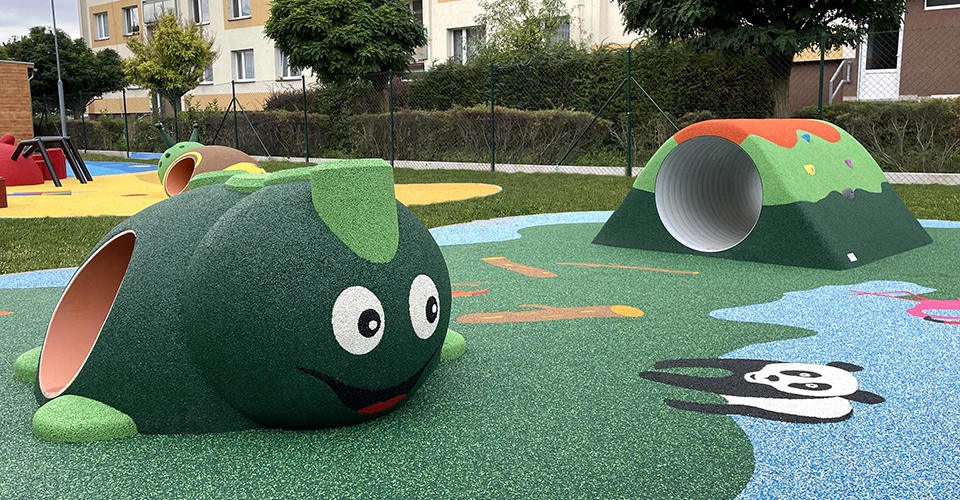 Children's playground featuring a green caterpillar tunnel, a small hill with a tunnel, and a panda illustration on the ground, with buildings and trees in the background.