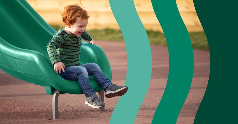 A young child with red hair wearing a green striped hoodie and jeans slides down a green playground slide on a sunny day.