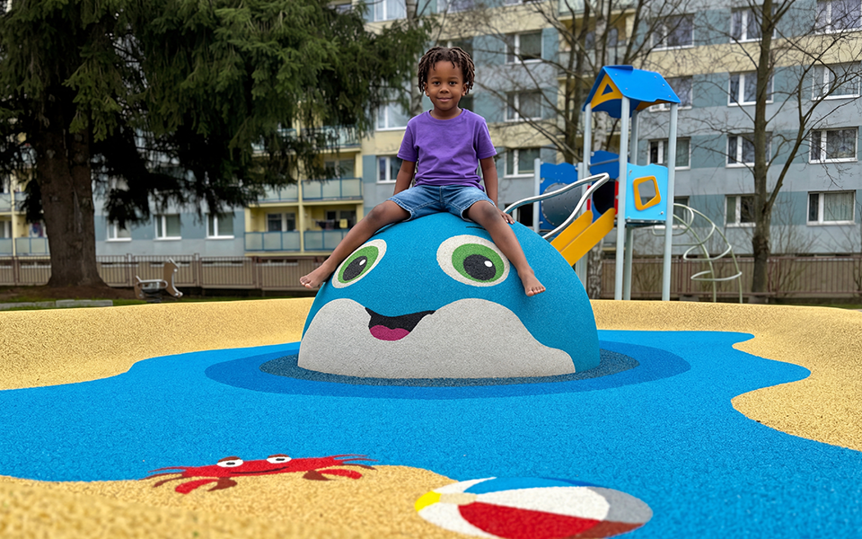 A child sits on a blue whale-shaped playground structure with a slide, surrounded by colorful designs including a crab and a beach ball. Apartment buildings and trees are in the background.