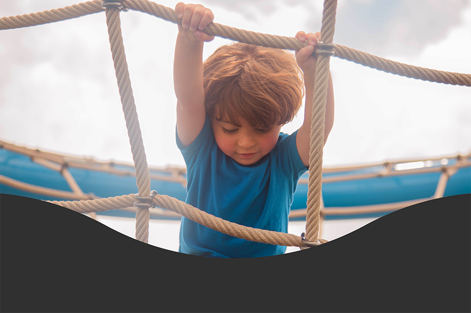 A young child in a blue shirt climbs a rope net outdoors, holding onto the ropes with both hands.