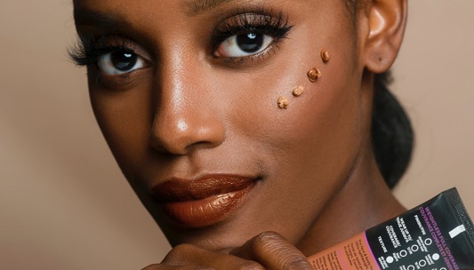 A woman with smooth brown skin applies several dots of liquid foundation to her cheek while holding a tube of makeup product.